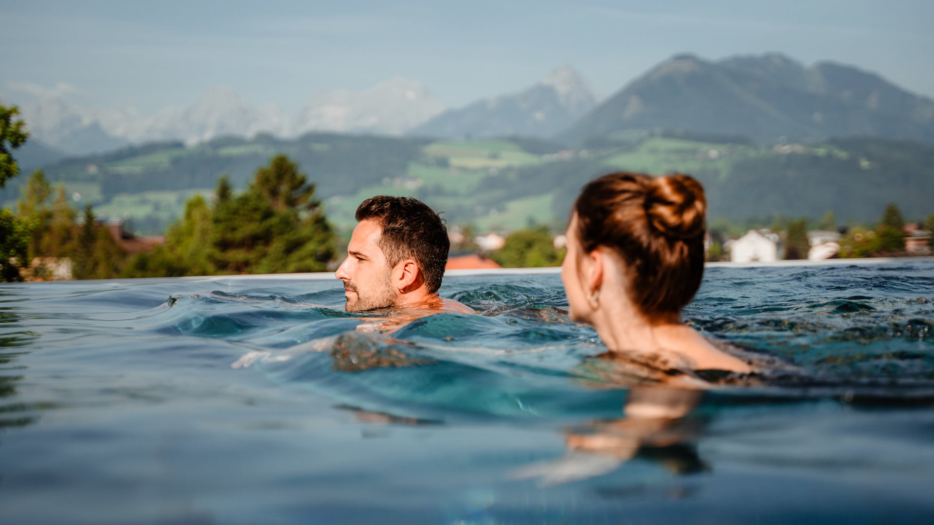 Mann und Frau schwimmen im Außenpool mit Blick auf die Berge im Wellnessurlaub in Oberösterreich